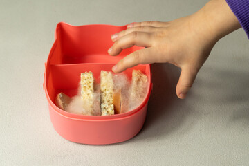 A child's hand reaches for a lunchbox with slices of bread and sausage on which white mold. Spoiled bad food in mold.