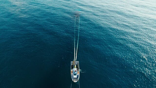 White small fisherman trawler with crew on board. People work together to pull out net with caught sardines from deep blue waters of open sea. Aerial shot with front view