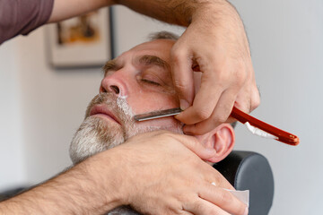 Barber trimming and cutting bearded man with shaving machine in barbershop. Hairstyling process.