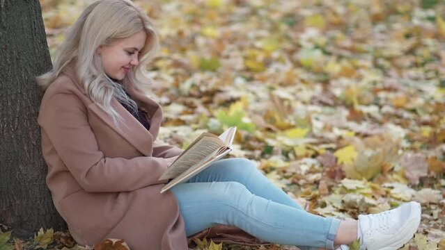 Enjoying reading. Smiling woman. Romantic fall. Pretty happy lady sitting on leaves ground leaning tree with interesting book slow motion.