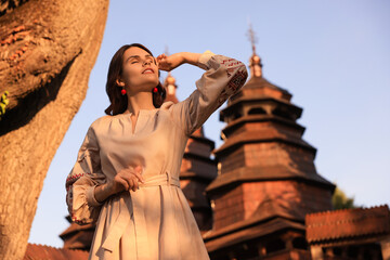 Fototapeta premium Beautiful woman wearing embroidered dress near old wooden church in village. Ukrainian national clothes