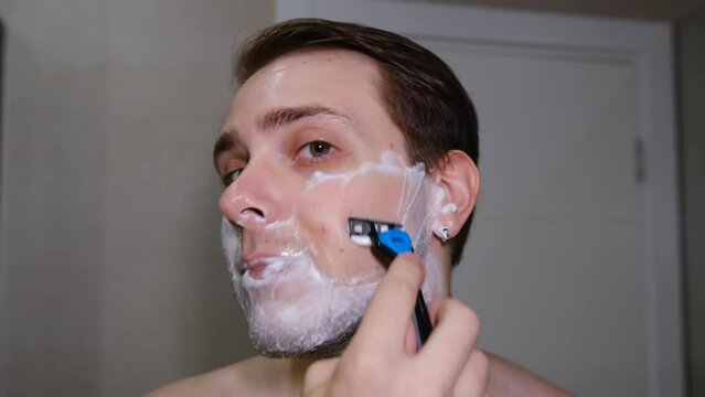 Serious young guy shaving his beard in a bathroom by razer.