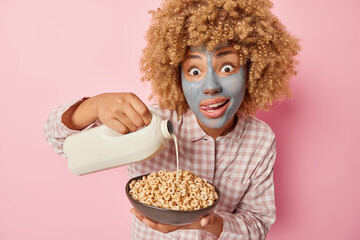 Surprised curly haired woman pours milk in cornflakes going to have tasty breakfast focused at...