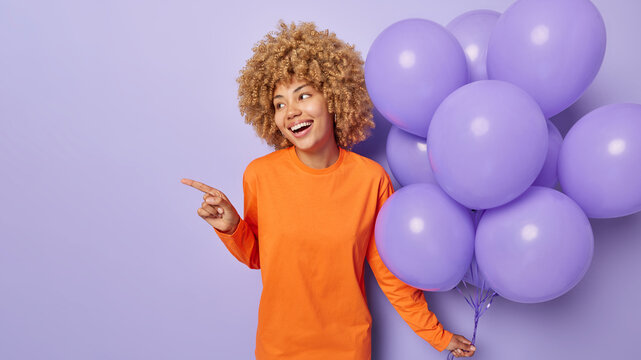 Studio shot of happy blonde woman smiles broadly dressed in casual orange jumper points index finger away holds bunch of inflated balloons celebrates special occasion isolated over purple background