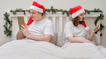 A woman and a man are looking at a tablet facing away from each other on a bed decorated for Christmas on New Year Eve, couple in a home bedroom