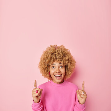 Smiling woman with curly blonde hair points fingers up shows way to advertisement demonstrates product has happy expression wears casual jumper isolated over pink background promots company.