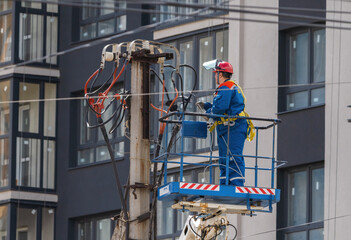 An electrician raised in the cradle of a tower lift mounts wires on a pole against the background...