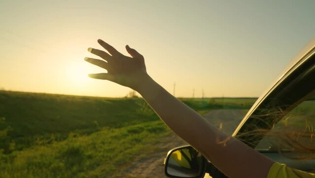 Young girl driver sits in front seat of car, sticking her hand out of window catching wind, glare of setting sun. Free woman travels by car, child travels by car. Teenage student travels by rental car