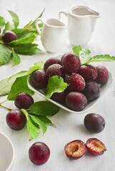 On a white wooden table is a small white dish with fresh red plums with leaves. Plums with leaves and white dishes are also on the table nearby. Vertical frame orientation.
