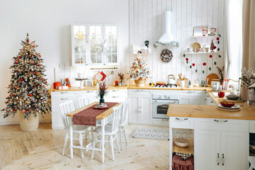 White Scandinavian style kitchen decorated for Christmas. Dining table and Christmas tree with garland lights and red and silver Christmas toys. Christmas interior. Empty space