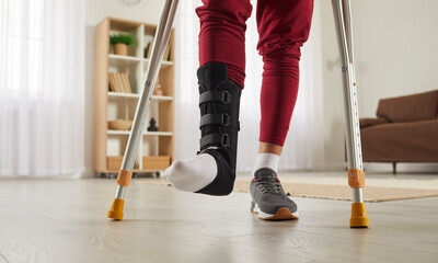 Man with broken leg or foot injury walking with crutches at home. Young guy wearing ankle support...