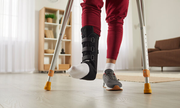 Man with broken leg or foot injury walking with crutches at home. Young guy wearing ankle support brace or fracture fixator with adjustable straps standing in living room. Cropped low section close up