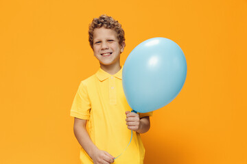 a cute, pleasant boy with curly hair stands on a yellow background holding a blue balloon in his hand