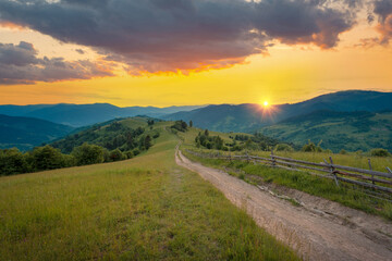 Naklejka na ściany i meble Dramatic colorful summer sunset sky over mountain hill with the rural road. Carpathian mountains. Ukraine.