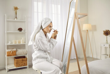 Beautiful young woman in a white dressing gown and bath towel turban on her hair sitting in front of a big mirror in the bedroom, looking at her reflection and applying facial beauty mask on her skin