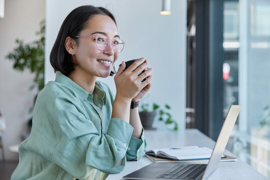 Horizontal shot of happy Asian woman with dark bob hair has coffee break while working at laptop computer poses against cozy interior watches education webinar connected to wireless internet