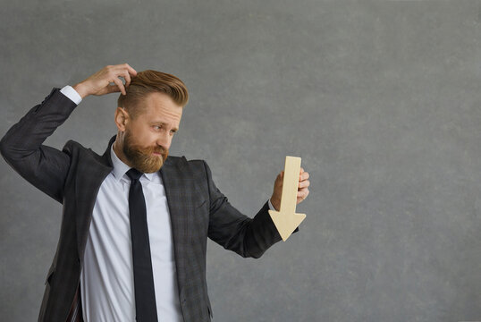 Confused businessman in suit holding down value fall arrow scratching head studio shot. Young adult puzzled caucasian entrepreneur thinking about recession. Achievement career wealth business