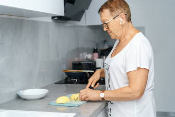 Senior Woman Cutting Potatoes On Kitchen Board, Wearing Earbud Headphones And Listening Music.