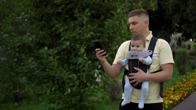 A young father walks in the park with a newborn child with a phone in his hands. The man is looking at the phone intently. Child in kangaroo-backpack. Closeup.