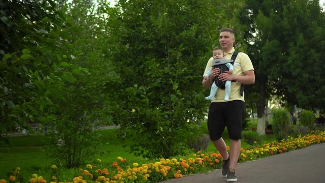 A young man walks along the alley in the park. A single father walks with his son. Newborn son in a kangaroo-backpack.