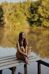 A young happy woman with a hippie smile sits on the lake shore on a bridge wearing eco clothing made of natural materials in harmony with nature in the fall. Travel lifestyle