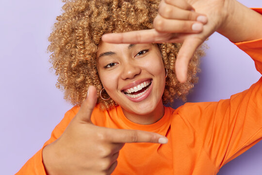 Cheerful curly haired adult woman makes frame gesture takes good shot smiles toothily captures moment searches perfect angle dressed in casual orange jumper isolated over purple studio background.