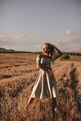 Stylish blonde girl with long curly hair in a light long dress stands in a field at sunset and looks away. 