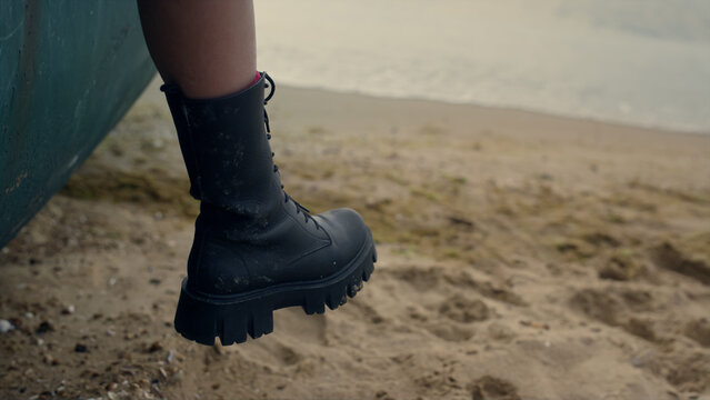 Woman leg wearing black boot hanging from old boat standing sand beach close up. - Powered by Adobe