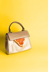 A small women's handbag in gold color with a slice of pepperoni pizza on a yellow background.  Creative concept of on-the-go snacking. Classic daily style.