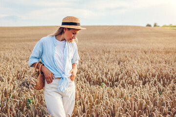 Young woman walking in summer field wearing straw hat and linen shirt holding handbag with wheat bundle. Space