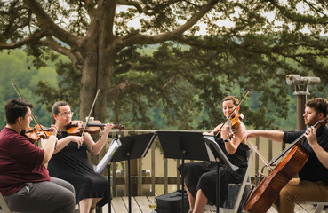 String quartet of two young men and two young women playing concert on wooden deck above Missouri River on summer evening; river and woods in background