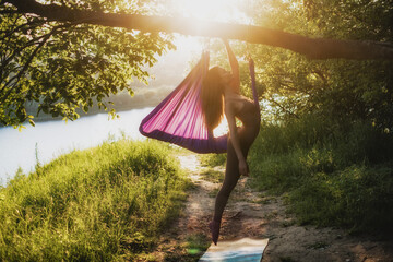 A young female gymnast is engaged in aerial yoga, using a combination of traditional yoga poses,...