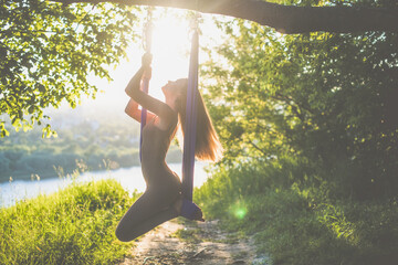 A young female gymnast is engaged in aerial yoga, using a combination of traditional yoga poses, pilates and dance using a hammock at sunset in nature. Healthy lifestyle. - obrazy, fototapety, plakaty