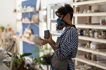 Ceramist woman in apron working with clay mug in ceramic workshop, wear protective respirator. Young creative potter in studio grinding crockery from raw crockery before baking. Small business, hobby.