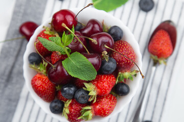 A white plate with cherries, strawberries and blueberries decorated with mint stands on a striped towel, on which lies a spoon, berries and mint leaves on a white background close-up. High quality