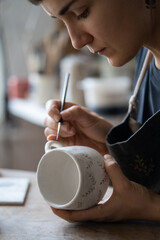 Fototapeta na wymiar Hands of professional artisan hold white ceramic mug drawing pattern with dark paint using brush on blurred background. Woman enjoys working with handmade pottery craft in studio extreme closeup
