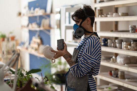 Ceramist woman in apron working with clay mug in ceramic workshop, wear protective respirator. Young creative potter in studio grinding crockery from raw crockery before baking. Small business, hobby.