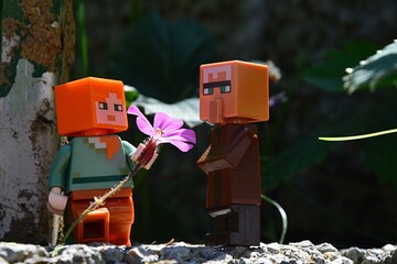 Fototapeta premium LEGO Minecraft figure of Alex handing pink chalice shaped flower to villager mob while standing on stone murral in real garden, summer daylight sunshine. 