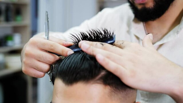 A barber man cuts a client's hair with scissors under a comb in a hairdressing salon.