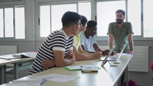 Young Professor helping multi ethnic male students in High School classroom. Friendly Teacher with beard explaining exercicse to latin, caucasian and african boys - Diversity in Education