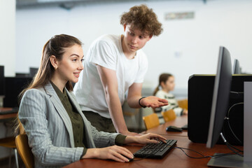 Interested teenage student talking to young female teacher sitting at computer in information technology class..