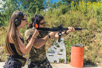 Fototapeta premium Woman standing next to man aiming submachine gun both wearing safety headphones and goggles. Firearms training at firing range. Outdoor horizontal shot. High quality photo