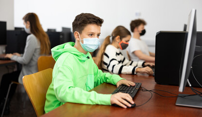 Diligent tween boy in protective face mask studying with classmates in modern computer lab at...