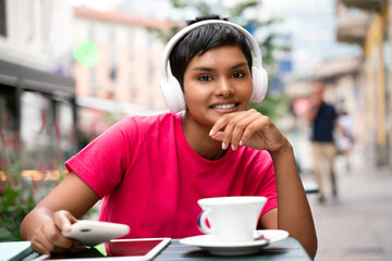 Portrait of smiling cheerful Indian woman listening music wearing wireless headphones, drinking coffee in cafe. Coffee break concept 