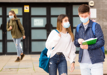 Focused teenage girl and boy in face masks talking about homework after lessons near school outdoors