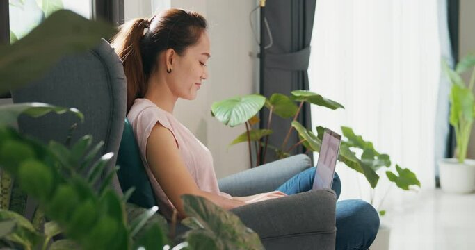 Young Asian woman wear casual calm sitting on wing chair use laptop computer thinking the idea for working full of plants in living room greenhouse. Coronavirus and stay home, Work from home concept.