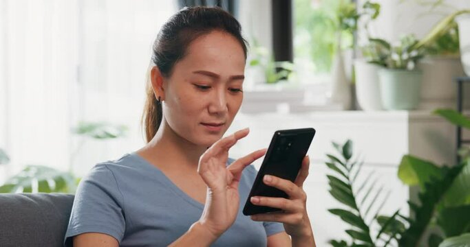 Close-up Young Asian woman wear casual site on couch use smartphone feel exciting scrolling screen online shopping in living room indoor plants at home. Excited and surprised female holding cellphone.