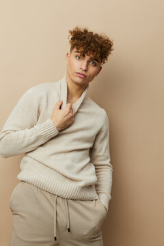  portrait of a handsome man with curly hair, standing on a beige background in a light turtleneck and looking at the camera with his hands in his pants pockets, while the other pulls back the collar