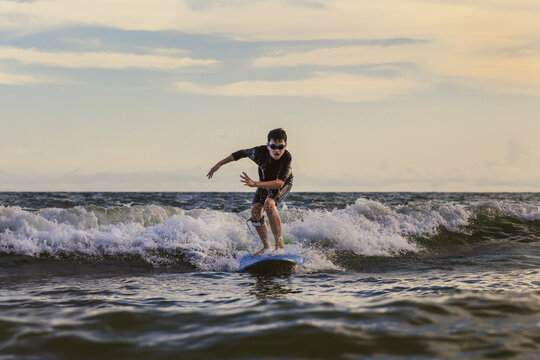 Young boy kid surfer riding waves with a soft board in Rayong beach, Thailand. Rookie surfboard student playing on water in excited face and funny action.