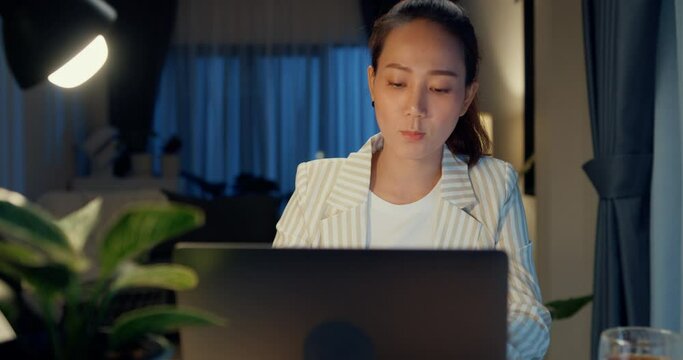 Young Asian businesswoman wear formal sit front of desk with laptop computer and cup of tea focus on screen check report meeting in cozy living room indoor plant at home night. Overtime work concept.
